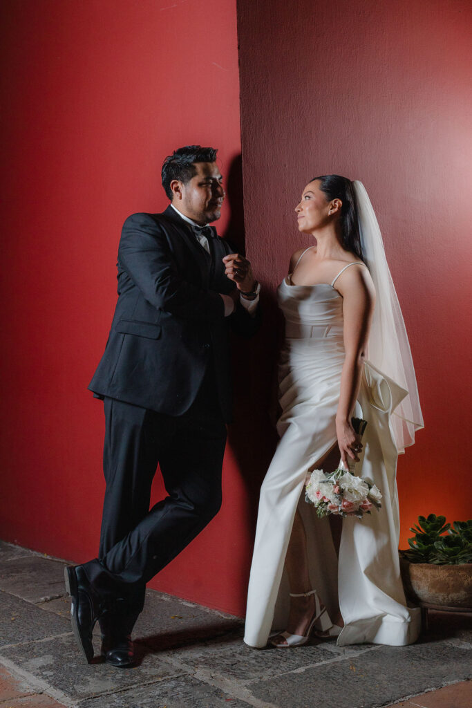 Novios mirándose frente a una pared roja durante una sesión de fotografía de bodas elegante. Retrato romántico con composición artística, estilo editorial y luz natural.