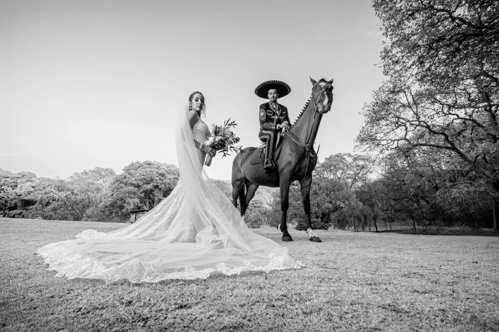 Fotografía editorial de bodas en blanco y negro de una novia y un novio vestido de charro sobre un caballo. El vestido de novia destaca en primer plano mientras el paisaje campestre aporta una composición elegante y atemporal.