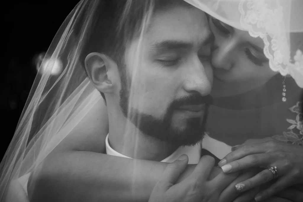 Fotografía artística de una pareja de novios bajo el velo de la novia. Retrato editorial de bodas que utiliza la transparencia del tul, la luz natural y una composición fine art para crear una imagen elegante, íntima y atemporal.