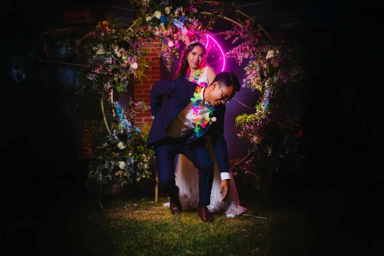 Novios posando de forma divertida bajo un arco floral iluminado con luces de neón durante la recepción de su boda, en un retrato artístico y elegante.