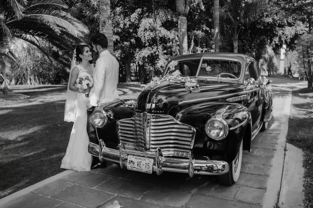 Novia con su ramo y novio recargado en un automóvil clásico durante una sesión de fotos de boda con un estilo elegante y atemporal.