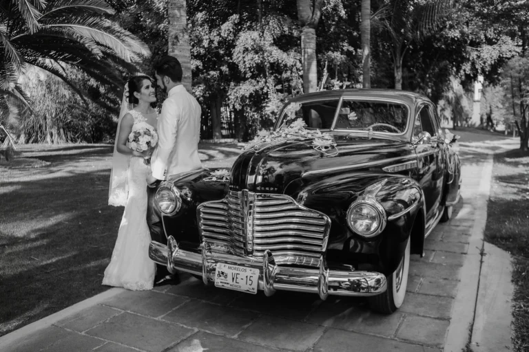 Novia con su ramo y novio recargado en un automóvil clásico durante una sesión de fotos de boda con un estilo elegante y atemporal.